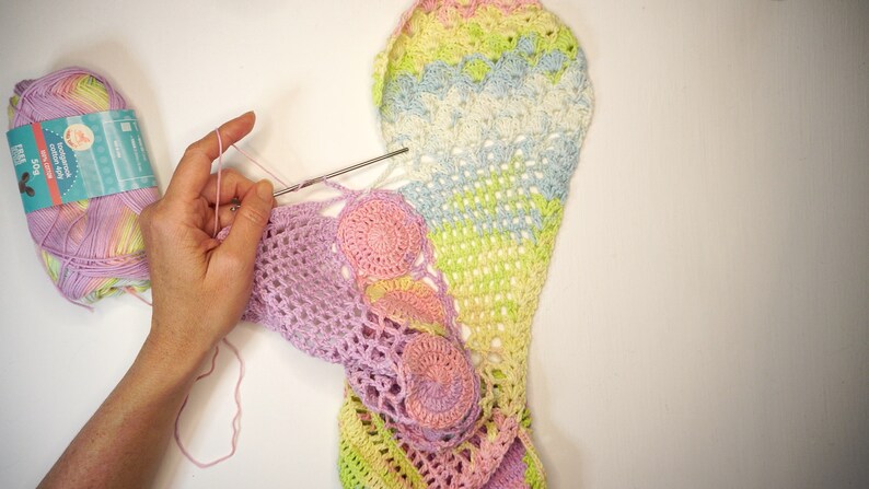 May include: A person is crocheting a colorful, multi-colored crochet blanket with a pink, green, and yellow color scheme. The blanket is made with a variety of stitches, including a crochet shell stitch and a crochet granny square stitch.