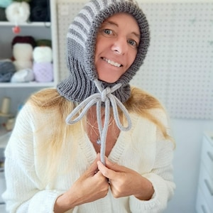 Könnte beinhalten: Ein gestrickter Kapuzen-Schal in Grau und Weiß mit einer Binde zum Verschließen. Der Schal wird von einer Person mit blonden Haaren getragen.