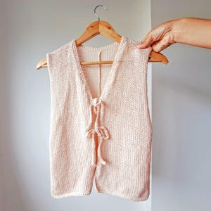 May include: A light pink knitted sleeveless vest with a tie closure at the neckline.