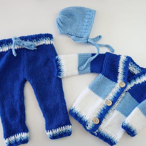 May include: A knitted baby outfit in shades of blue and white. The set includes pants with a drawstring, a button-up cardigan, and a bonnet. The cardigan has wooden buttons.