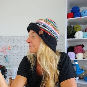 Op de afbeelding: Een vrouw draagt een zwarte en meerkleurige gestreepte gebreide beanie met een zwarte rand. De beanie heeft een bruin leren label met het woord "Wool" erop.