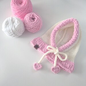 Könnte beinhalten: Eine handgefertigte, weiß-rosa gestrickte Babyhaube mit Schleife und herzförmigen Bommeln. Drei Knäuel Wolle in Weiß und Rosa liegen neben der Haube. Die Haube hat ein kleines braunes Etikett.