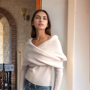 May include: A white knitted wrap with long sleeves, worn over a white tank top and blue jeans.