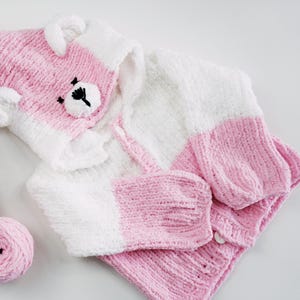 以下が含まれることがあります： ピンクと白のニットベビー服セット。クマの耳が付いたフード付きジャケットと、お揃いのミトンが含まれています。クマの顔はフードに刺繍されています。ピンクの毛糸玉も見えます。