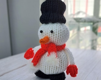 May include: A handmade knitted snowman with a black top hat, orange carrot nose, and red scarf and mittens. The snowman has black button eyes and a white pom-pom belly. The snowman is a winter holiday decoration.