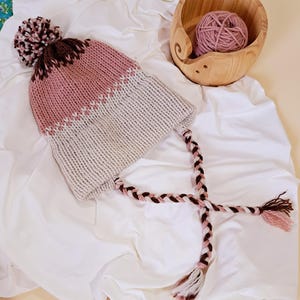 May include: A hand-knitted winter hat in shades of pink, brown, and cream, featuring a pom-pom and braided ties. The hat is made of soft yarn and is designed for warmth and style. A wooden bowl with a ball of yarn is also in the image.