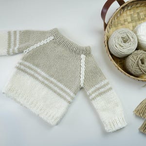 May include: A handmade, knitted sweater in neutral tones of beige and white. The sweater features a striped design on the lower half and sleeves. A basket with yarn balls and decorative tassels are in the background.