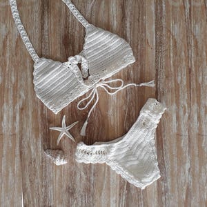 May include: White crochet bikini top and bottom with a tie closure on the top. The bikini is laid out on a wooden surface.