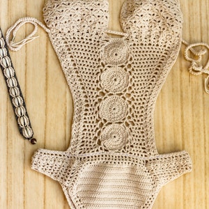May include: A beige crochet swimsuit with a cutout design and a decorative circle pattern. The swimsuit is laid flat on a wooden surface.