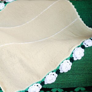 Puede incluir: Una manta de bebé de punto de color crema con un borde de ganchillo blanco con flores blancas y hojas verdes.