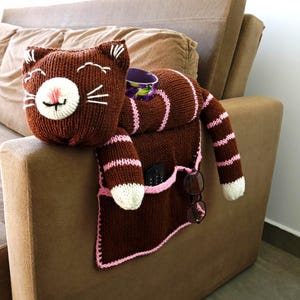 Pode incluir: Um organizador de apoio de braço de sofá em forma de gato castanho tricotado com detalhes em rosa. O gato tem focinho e bigodes brancos, e o organizador tem bolsos para um controlo remoto e óculos. Um recipiente roxo é visível.