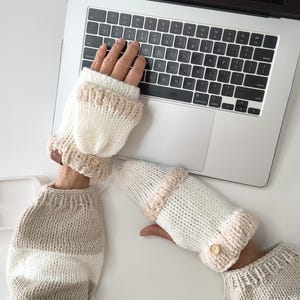 Könnte beinhalten: Weiß gestrickte Fingerhandschuhe mit beigem Besatz und Holzknopf. Die Handschuhe werden beim Tippen auf einem Laptop getragen. Die Ärmel sind beige und weiß gestreift. Warme Accessoires für den Winter.