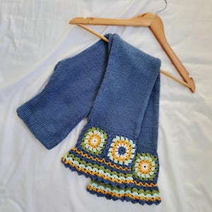 May include: A blue knitted scarf with a crocheted trim featuring colorful flowers. The scarf is hanging on a wooden hanger.