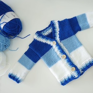Peut inclure: Un cardigan tricoté à la main, rayé bleu, blanc et bleu clair, avec des boutons en bois. Le cardigan est présenté avec des pelotes de laine bleues et blanches, suggérant un projet artisanal. Le vêtement est conçu pour un bébé ou un jeune enfant.