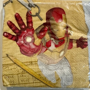May include: A brown and yellow paper napkin with a red and gold illustration of Iron Man. The napkin has a grid pattern and the text "STARK INDUSTRIES" is visible in the bottom right corner.