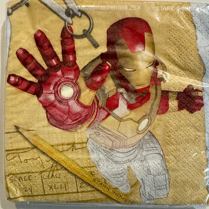 May include: A brown and yellow paper napkin with a red and gold illustration of Iron Man. The napkin has a grid pattern and the text "STARK INDUSTRIES" is visible in the bottom right corner.