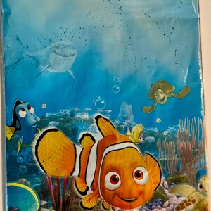 Puede incluir: Un mantel con el tema de Buscando a Nemo. El diseño presenta a Nemo, Dory y otros personajes de la película sobre un fondo submarino. El mantel mide 137 x 259 cm.
