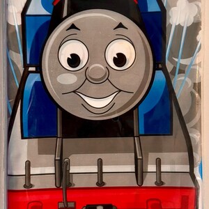 Könnte beinhalten: Eine blaue und rote Cartoon-Lokomotive mit einem lächelnden Gesicht und großen Augen. Der Zug befindet sich auf einem Gleis mit braunen Holzschwellen. Der Hintergrund ist ein blaues und weißes Muster mit Sternen.