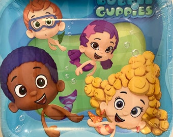 Puede incluir: Un plato de papel azul y blanco con una ilustración colorida de los personajes de Bubble Guppies. Los personajes están nadando en una escena de océano azul y verde. El texto "Bubble Guppies" está impreso en letras blancas sobre un fondo azul.