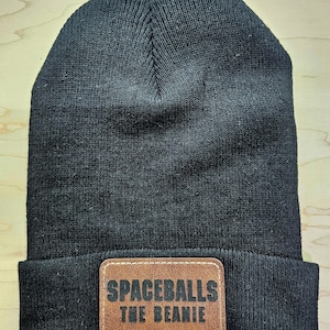 May include: Black knit beanie with a folded brim. A brown leather patch on the front reads "SPACEBALLS THE BEANIE" in dark lettering. The hat is made of a soft, textured material.