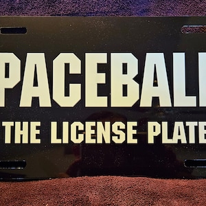 May include: Black license plate with the words "SPACEBALLS" in large white block letters above "THE LICENSE PLATE". The plate has four mounting holes and a glossy finish.