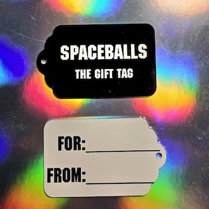 May include: Two gift tags. The top tag is black with the text "SPACEBALLS THE GIFT TAG" in white. The bottom tag is white with the text "FOR:" and "FROM:" in black. Both tags have a scalloped edge and a hole for attaching.