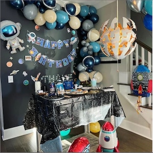 May include: A space-themed birthday party setup with a table covered in a black, glittery tablecloth. Decorations include balloons in shades of blue, gray, and white, a "Happy Birthday" banner, and astronaut and rocket ship balloons. A rocket ship pi&ntilde;ata is also present.