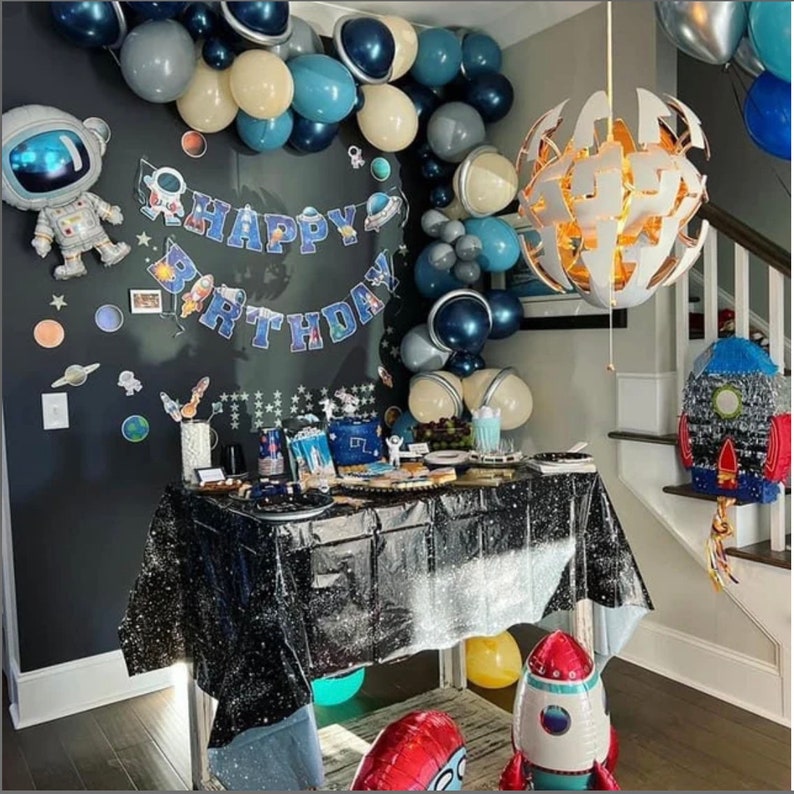 May include: A space-themed birthday party setup with a table covered in a black, glittery tablecloth. Decorations include balloons in shades of blue, gray, and white, a "Happy Birthday" banner, and astronaut and rocket ship balloons. A rocket ship pi&ntilde;ata is also present.