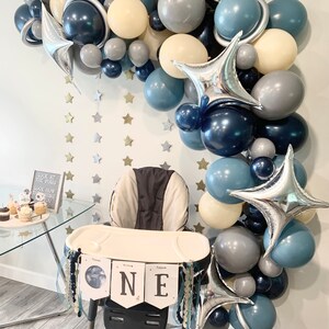 May include: A space-themed first birthday party setup. A high chair is decorated with a banner that says "ONE". A balloon arch in shades of blue, gray, and cream, with star-shaped foil balloons and star cutouts on the wall.
