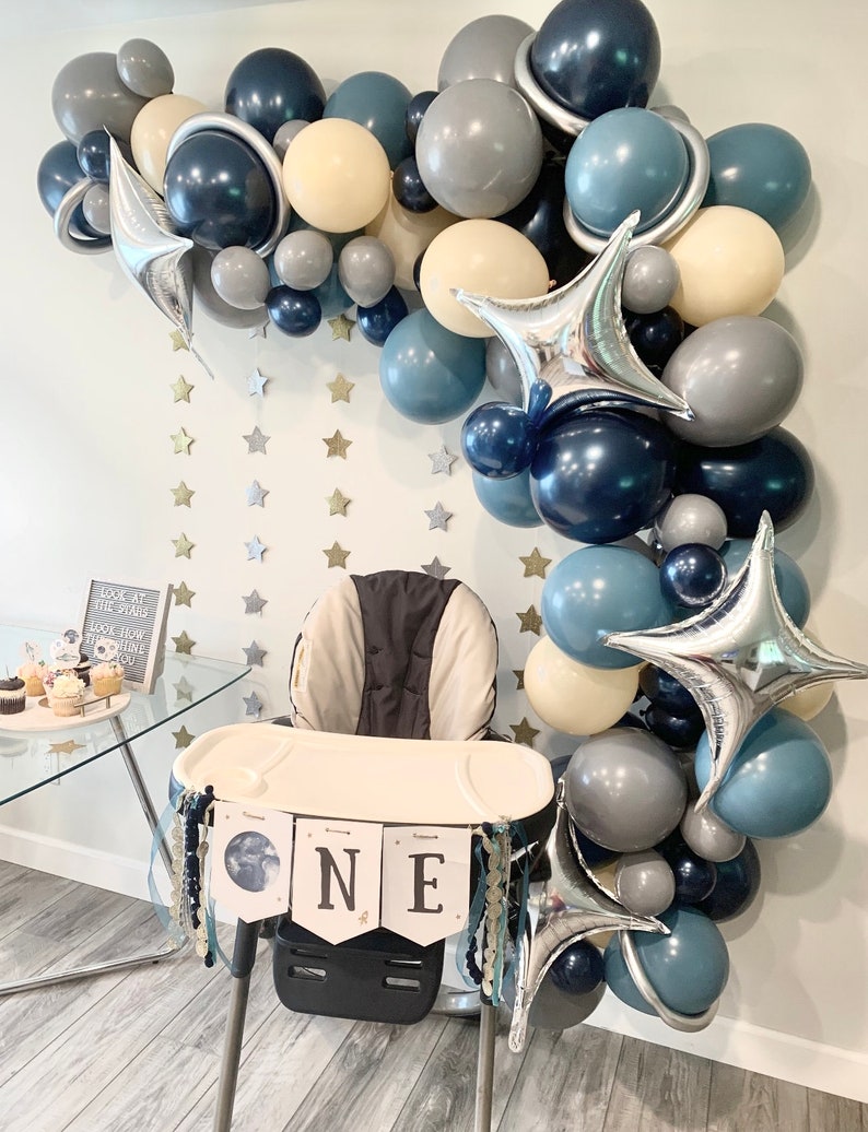 May include: A space-themed first birthday party setup. A high chair is decorated with a banner that says "ONE". A balloon arch in shades of blue, gray, and cream, with star-shaped foil balloons and star cutouts on the wall.