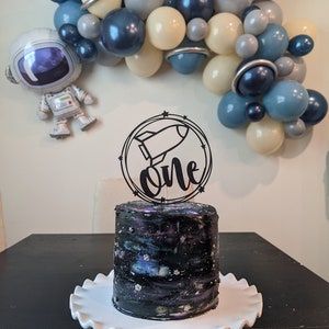 May include: A birthday cake with a galaxy design, topped with a black rocket ship and the word "one". The cake sits on a white, ruffled cake stand. A balloon arch in shades of blue, gray, and cream, and an astronaut balloon decorate the background.