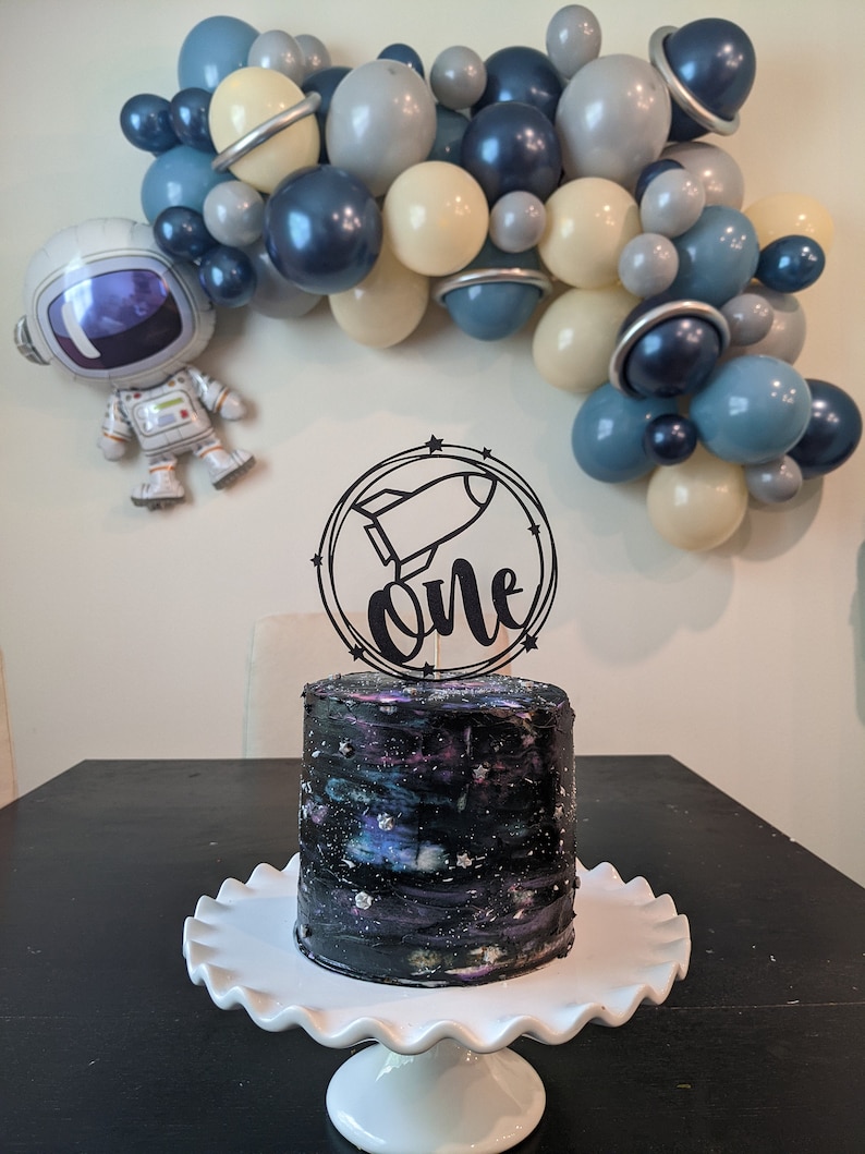 May include: A birthday cake with a galaxy design, topped with a black rocket ship and the word "one". The cake sits on a white, ruffled cake stand. A balloon arch in shades of blue, gray, and cream, and an astronaut balloon decorate the background.