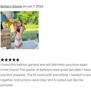 May include: A woman holding a baby stands near a balloon garland with gold star and blue, white, and gold balloons. The woman wears a striped dress. The garland is for a party.