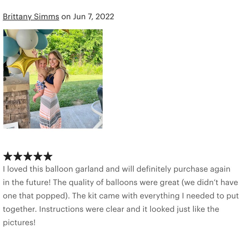 May include: A woman holding a baby stands near a balloon garland with gold star and blue, white, and gold balloons. The woman wears a striped dress. The garland is for a party.