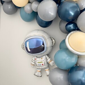 May include: An astronaut-shaped foil balloon with a reflective visor and white spacesuit is featured. The balloon is surrounded by a balloon arch in shades of blue, gray, and cream, with planet-shaped balloons.