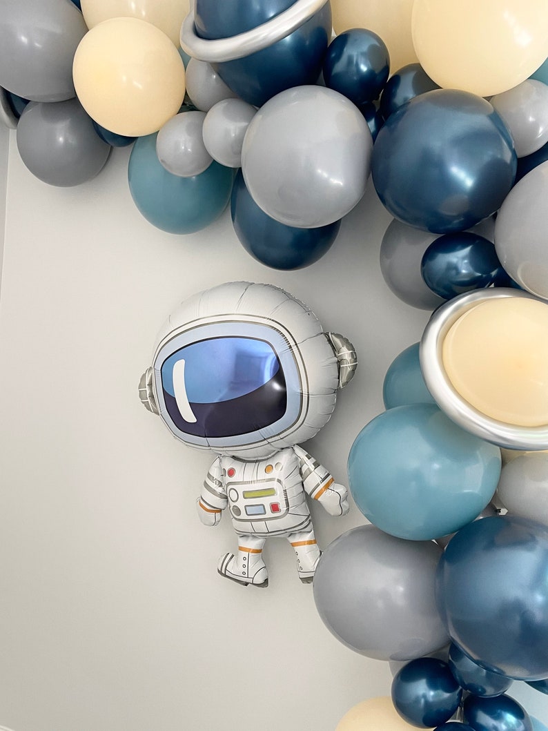 May include: An astronaut-shaped foil balloon with a reflective visor and white spacesuit is featured. The balloon is surrounded by a balloon arch in shades of blue, gray, and cream, with planet-shaped balloons.
