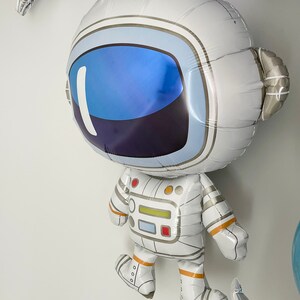 May include: A large, white astronaut-shaped balloon with a blue visor and silver accents. The balloon has detailed features, including a helmet, spacesuit, and boots. Other star-shaped balloons are visible in the background, creating a space-themed party atmosphere.