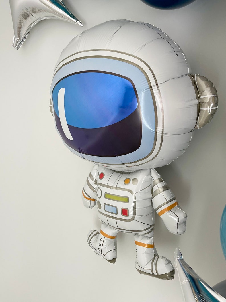 May include: A large, white astronaut-shaped balloon with a blue visor and silver accents. The balloon has detailed features, including a helmet, spacesuit, and boots. Other star-shaped balloons are visible in the background, creating a space-themed party atmosphere.