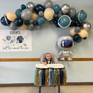 May include: A festive party scene with a balloon arch in shades of blue, gray, and beige. A banner reads "Elijah's first trip around the sun." An astronaut balloon and a high chair with tassel decorations complete the space-themed decor.