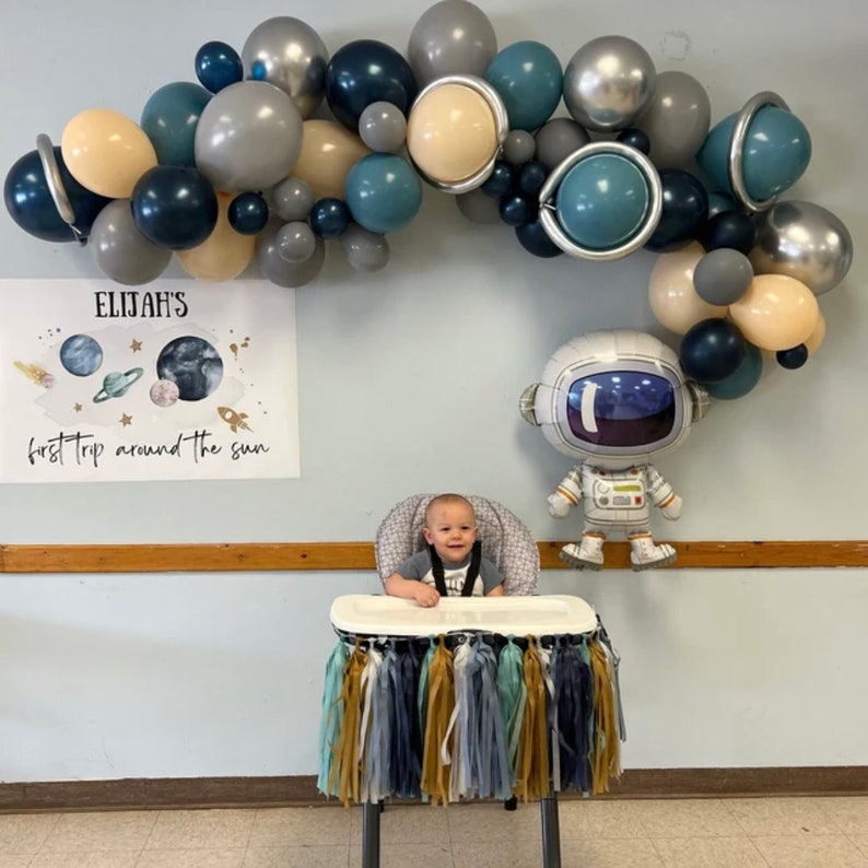 May include: A festive party scene with a balloon arch in shades of blue, gray, and beige. A banner reads "Elijah's first trip around the sun." An astronaut balloon and a high chair with tassel decorations complete the space-themed decor.
