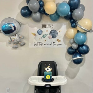 May include: A space-themed birthday party setup with a cake, balloons, and a banner. The cake is decorated with a rocket and the number 1. Balloons in shades of blue, gray, and cream form an arch. A foil astronaut balloon is also present.
