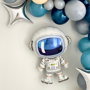 May include: An astronaut-shaped foil balloon with a white spacesuit and a reflective blue visor. The balloon is part of a space-themed balloon arch, featuring various shades of blue, gray, and cream-colored balloons, along with silver star-shaped foil balloons.