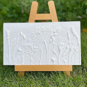 May include: A white rectangular plaque with a raised relief design of pressed wildflowers and grasses. The plaque is displayed on a small wooden easel.