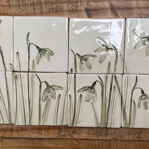 May include: A collection of square ceramic tiles featuring hand-painted green snowdrop flowers on a glossy white background. The tiles are arranged in two rows, displaying various botanical illustrations of drooping white petals and long green stems.