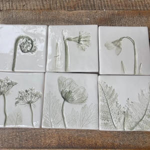 May include: Six square ceramic tiles with botanical designs. Each tile features a different embossed plant, including a fern, daffodil, and poppy, in shades of green and white. The tiles are arranged on a wooden surface.