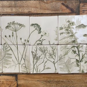 May include: A set of ten square ceramic tiles arranged in two rows of five. Each off-white tile features a detailed, embossed botanical print in a muted sage green colour, depicting various wildflowers, ferns, and grasses with intricate leaf textures.