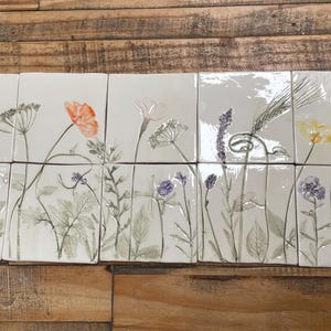 May include: A set of ten square white ceramic tiles arranged in two rows, each featuring detailed botanical impressions of wildflowers, grasses, and ferns. The designs include delicate stems, leaves, and colourful floral accents in orange, yellow, and purple.