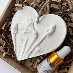 May include: White ceramic heart-shaped ornament with a raised floral design. The ornament has a hole at the top for hanging. A small glass bottle with a gold cap and a white label is included.