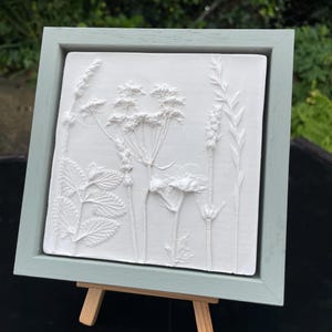May include: A white relief artwork featuring botanical elements, framed in a square, pale blue-green wooden frame. The piece is displayed on a small wooden easel. The background is blurred, suggesting an outdoor setting. The artwork showcases detailed plant designs.