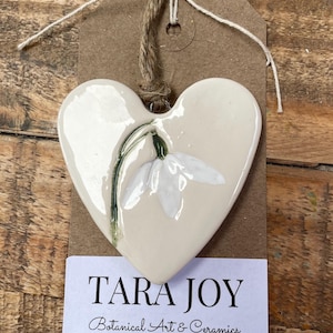 May include: Cream ceramic heart-shaped ornament featuring a white snowdrop flower design. The ornament hangs from a jute string and is attached to a brown tag with the text "TARA JOY Botanical Art & Ceramics".